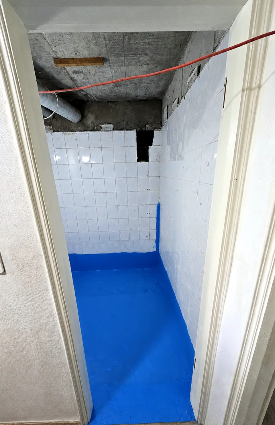 문 앞에서 본 방수 시공. 바닥 전체와 벽 아래쪽에 파란 방수층을 올렸고, 천장은 뜯어 콘크리트와 배관이 보인다